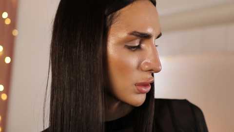 Video of a Middle Eastern woman with long, straight black hair, wearing a black top, looking down in thought. Background features warm, blurred lighting and a string of fairy lights.