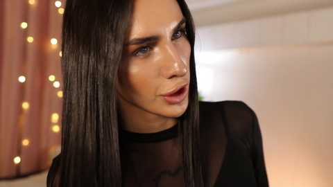 Video of a light-skinned woman with long, straight black hair, wearing a sheer black top, with a subtle golden glow in the background, creating a soft, cozy ambiance.