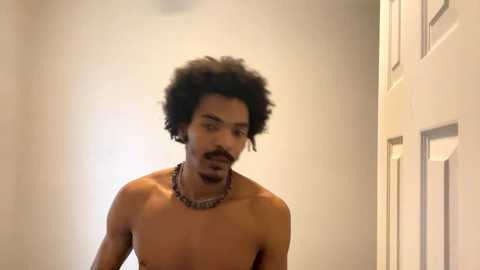 Media: Video of a shirtless Black man with a full afro and mustache, wearing a silver chain necklace, standing in a beige hallway with a door to the right.