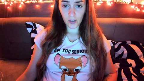 Media: Video of a young woman with long brown hair, wearing a white T-shirt with a cartoon mouse graphic and the text \"Are You Serious?\" in a cozy living room with string lights and a dark sofa.