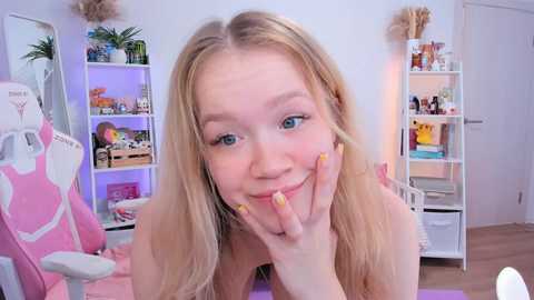 Media: Video of a young, light-skinned, blonde woman with blue eyes, smiling, in a brightly lit, modern bedroom. She has long nails painted yellow. Pink gaming chair in background.
