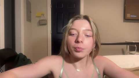 Media: Video of a young blonde woman with light skin and minimal makeup, wearing a green spaghetti-strap top, sitting on a bed in a dimly lit room with beige walls and a closed door.