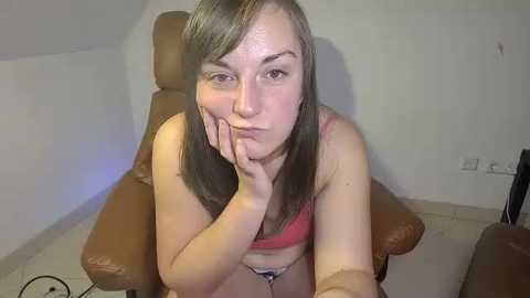 Media: Video of a fair-skinned woman with long brown hair, wearing a red bra, leaning on a brown leather chair, resting her chin on her hand, in a sparsely furnished room.