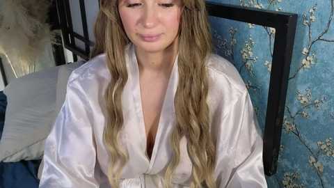 Media: A video of a young woman with long, wavy blonde hair wearing a silky, light pink robe, seated in a bedroom with blue floral wallpaper and a black-framed mirror.