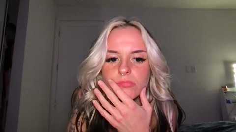 Media: Video of a young, fair-skinned woman with long, wavy platinum blonde hair, wearing a black top, and a septum piercing. She has her hand to her mouth, looking introspective. Background shows a simple, dimly-lit bedroom with a white door and a bed.