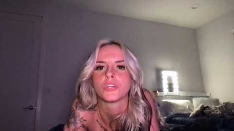 Media: Video of a young blonde woman with wavy hair, wearing a patterned top, standing indoors with dim lighting, white walls, and a partially visible bed in the background.