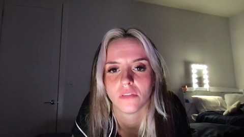 Media: A video of a young woman with long, platinum blonde hair and light skin, wearing a black top, in a dimly lit bedroom with a lit vanity mirror, a bed, and a closed door.