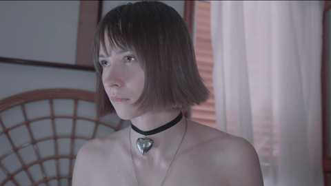 Media: Video of a young, pale-skinned woman with a short bob haircut and a black choker necklace featuring a heart pendant. She has a neutral expression and is seen from the shoulders up in a dimly lit room with wooden furniture and a window with white curtains.