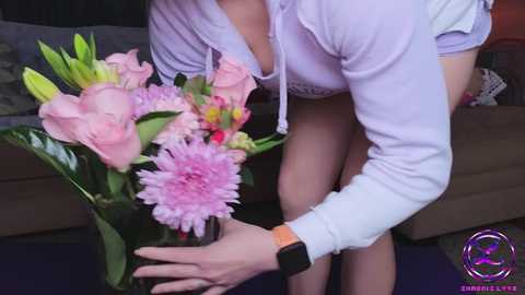 Media: Video of a woman in a white hoodie, holding a vase of pink flowers, with a wristwatch on her left wrist, standing in a living room.