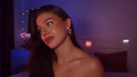 Media: Video of a young woman with long dark hair, wearing silver earrings, gazing thoughtfully. Background features a dimly lit room with purple and blue lighting, a bed, and a decorative lamp.