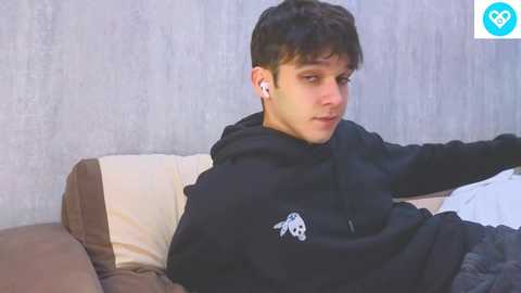 Media: A video of a young man with dark hair, wearing a black hoodie, lying on a beige and brown sofa, listening to white earbuds, in a simple, dimly lit room.