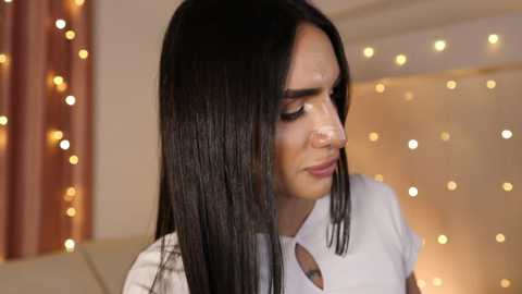 Video of a woman with straight, dark brown hair, fair skin, and a light pink lip color, wearing a white blouse, in a softly lit room with warm, bokeh lights and beige curtains.