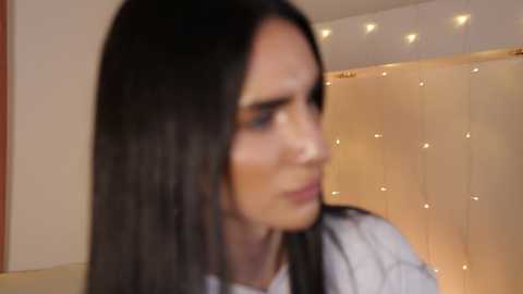 Video of a woman with long, straight black hair, fair skin, and light makeup, wearing a white top, against a warm-toned background with string lights. She appears contemplative.