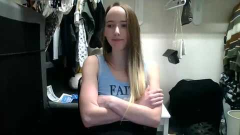 Media: Video of a young woman with long blonde hair, wearing a light blue tank top with \"FAU\" written on it, sitting in a cluttered closet with various clothes hanging and a white chair in the background.