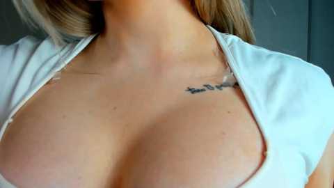 Media: Video of a light-skinned woman with long blonde hair, wearing a white top with a deep V-neck, showcasing her cleavage and a small tattoo on her collarbone.