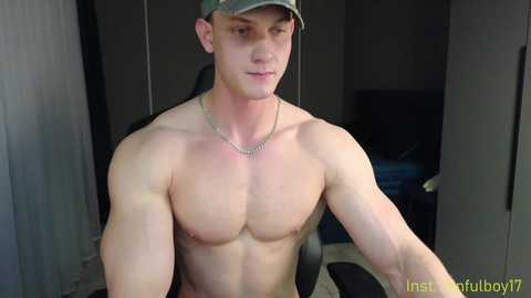 Media: Video of a shirtless, muscular white man with fair skin, wearing a green cap, sitting indoors. He has a silver chain necklace. Background shows a dark room with a chair and a TV.