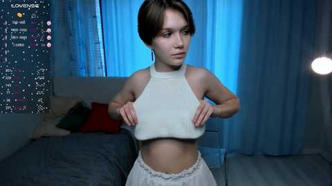 Media: A video of a young woman in a white crop top and pants, standing in a dimly lit bedroom with blue curtains, a bed, and a spotlight.