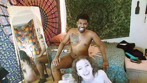 Media: Video of a naked, muscular Black man with a tattoo on his chest, smiling, sitting on a bed covered with a colorful tapestry. A woman with white face paint is standing beside him.