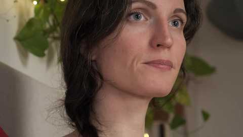 Media: Video of a fair-skinned woman with shoulder-length dark brown hair, blue eyes, and a slight smile, indoors with blurred green plants and a white wall in the background.