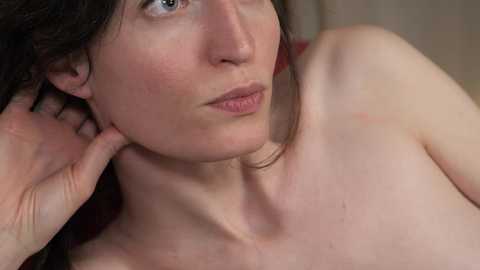 Media: Video of a fair-skinned, topless woman with long, dark hair, blue eyes, and freckles, holding her chin, against a blurred, neutral background.