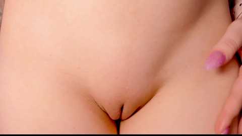 Media: A close-up video of a nude, light-skinned woman with smooth skin and visible pubic hair. Her hand, with pink nail polish, is resting on her thigh. The background is blurred, focusing attention on her genital area.