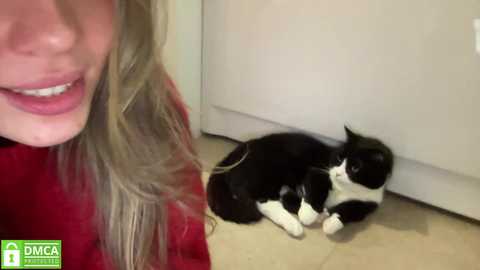 Media: Video of a smiling woman with wavy blonde hair, wearing a red sweater, sitting on a beige tiled floor. A black and white cat with a white chest and paws is lying beside her.