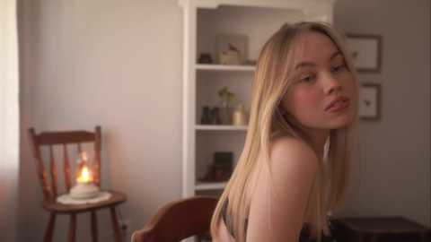 Media: Video of a young Caucasian woman with long blonde hair, wearing a black lace bra, sitting in a dimly lit room with a lit candle on a wooden table and white shelves in the background.