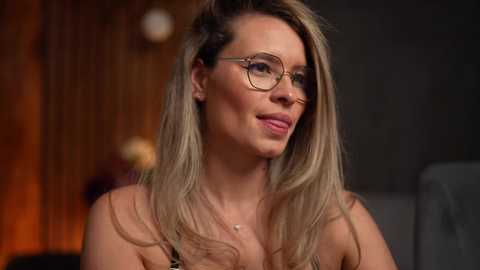 Media: Video of a smiling, fair-skinned woman with long blonde hair, wearing round glasses and a black top, seated in a dimly lit, modern room with wooden accents and a blurred, warm-toned background.