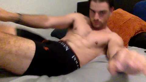 Media: Video of a shirtless, muscular man in black Calvin Klein underwear, lying on a bed with gray sheets and orange and blue pillows, looking directly at the camera.