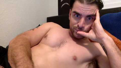 Media: A video of a shirtless, muscular man with a beard, blue eyes, and short brown hair, lying on a bed with a blue pillow. He is touching his chin with his left hand.