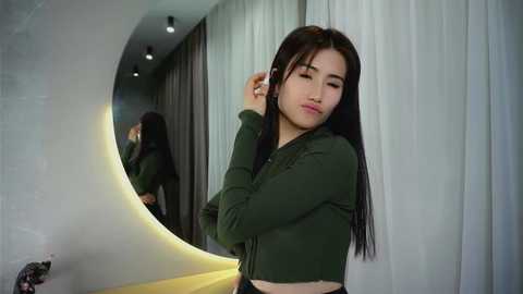 Media: Video of an East Asian woman with long black hair, wearing a green cropped sweater, adjusting her hair in a modern, minimalist room with a curved mirror.