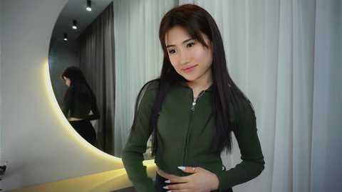 Media: Video of a young Asian woman with long black hair, wearing a dark green zip-up top, standing in front of a mirror with a curved, illuminated wall.