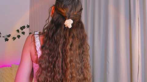 Media: Video of a woman's long, wavy brown hair styled with a white scrunchie, wearing a white lace bra, in a softly lit bedroom with pastel curtains and green garland.