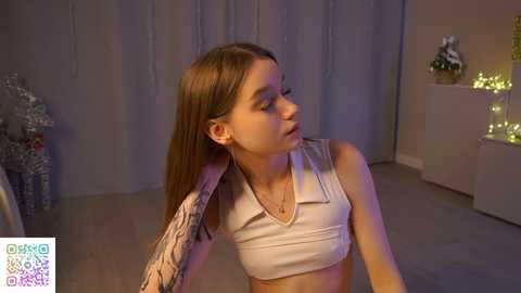 Media: Video of a young Caucasian woman with light skin, long brown hair, and a sleeve tattoo, wearing a white crop top, sitting indoors with a festive, lit-up background.