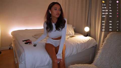 Media: Video of a young woman with long brown hair, wearing a white crop top and high-waisted shorts, sitting on a bed with white sheets and a fluffy pillow. A soft lamp and phone are nearby.