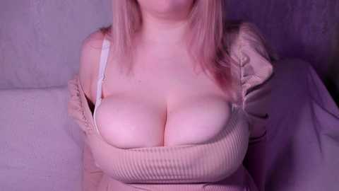 Media: Video of a fair-skinned woman with long, pink hair, wearing a beige, ribbed, low-cut top that accentuates her large breasts. She sits on a white couch against a textured, light gray wall. The image has a soft, purple-hued lighting.