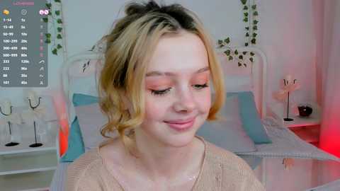 Media: Video of a smiling young woman with light skin, blonde hair, wearing light makeup and a beige knit sweater, standing in a cozy, pastel-colored bedroom with a white bed, green wall decorations, and soft lighting.