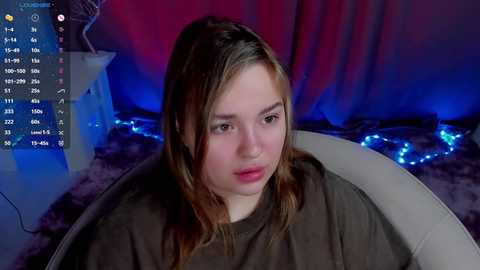 Media: Video of a young Caucasian woman with light skin and long brown hair, wearing a brown shirt, sitting in a white chair. Background includes a red curtain, blue fairy lights, and a white table.