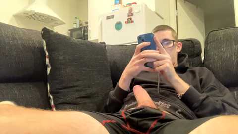 Media: Video of a young man with glasses and short hair, wearing glasses and black hoodie, holding a smartphone, seated on a dark couch in a modern kitchen with a white fridge.