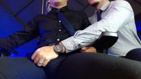 Media: Video of two men, one in black, the other in light gray, seated closely, the latter gripping the former's crotch, dimly lit background.