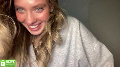 Media: Video of a smiling, light-skinned woman with long, wavy blonde hair, wearing a gray bathrobe. She has blue eyes and a cheerful expression.