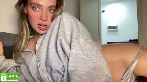 Media: Video of a blonde woman with fair skin, wearing a gray hoodie, sitting on a bed. She has a surprised expression, looking towards the camera. The background shows a white door and a bathroom with a towel rack.