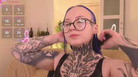 Video of a fair-skinned woman with blue hair, wearing glasses, black tank top, and intricate black and grey tattoos on her neck and arms, adjusting her hair in a modern, well-lit kitchen.