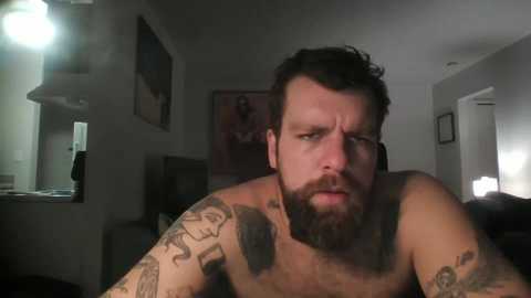 Media: Video of a bearded, muscular man with tattoos, looking serious, in a dimly-lit bedroom with a mirror and framed picture on the wall.