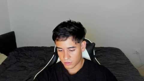 Media: Video of a young man with short black hair, wearing a black jacket and headphones, sitting on a gray bed with a dark quilt, in a minimalist room with a plain white wall.