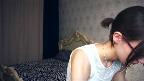 Media: Video of an Asian woman with dark hair tied back, wearing glasses and a white sleeveless top, standing in a room with zebra-patterned bedding, a wooden headboard, and gray wallpaper.