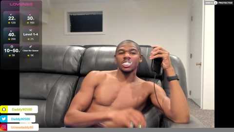 Media: Video of a shirtless Black man with a fit physique, wearing a black wristwatch, and holding a gaming controller, sitting on a grey leather couch in a modern living room.
