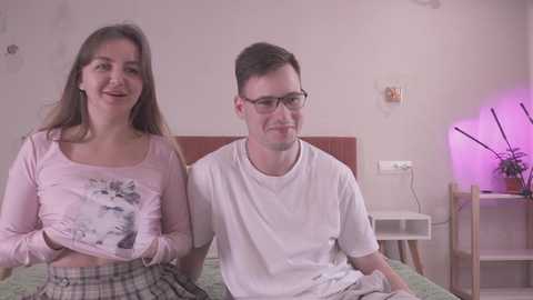 Media: Video of a young couple in a bedroom: woman with long brown hair in pink shirt, man with glasses in white shirt.
