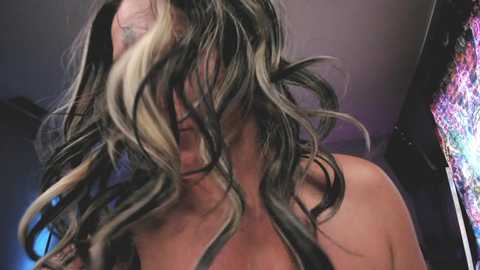 Media: A video of a woman with long, wavy black hair with blonde streaks, partially covering her face, wearing a strapless top, in a dimly lit room with purple and blue lighting and a colorful mural in the background.