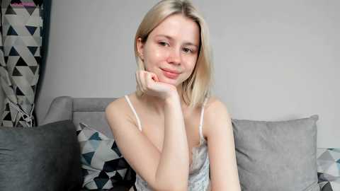 Media: Video of a young, light-skinned, blonde woman with straight hair, wearing a white camisole, resting her chin on her hand, sitting on a gray sofa with geometric-patterned pillows.